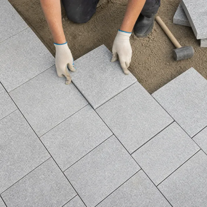 Hände in Arbeitshandschuhen verlegen hellgraue Granitplatten im Sandbett